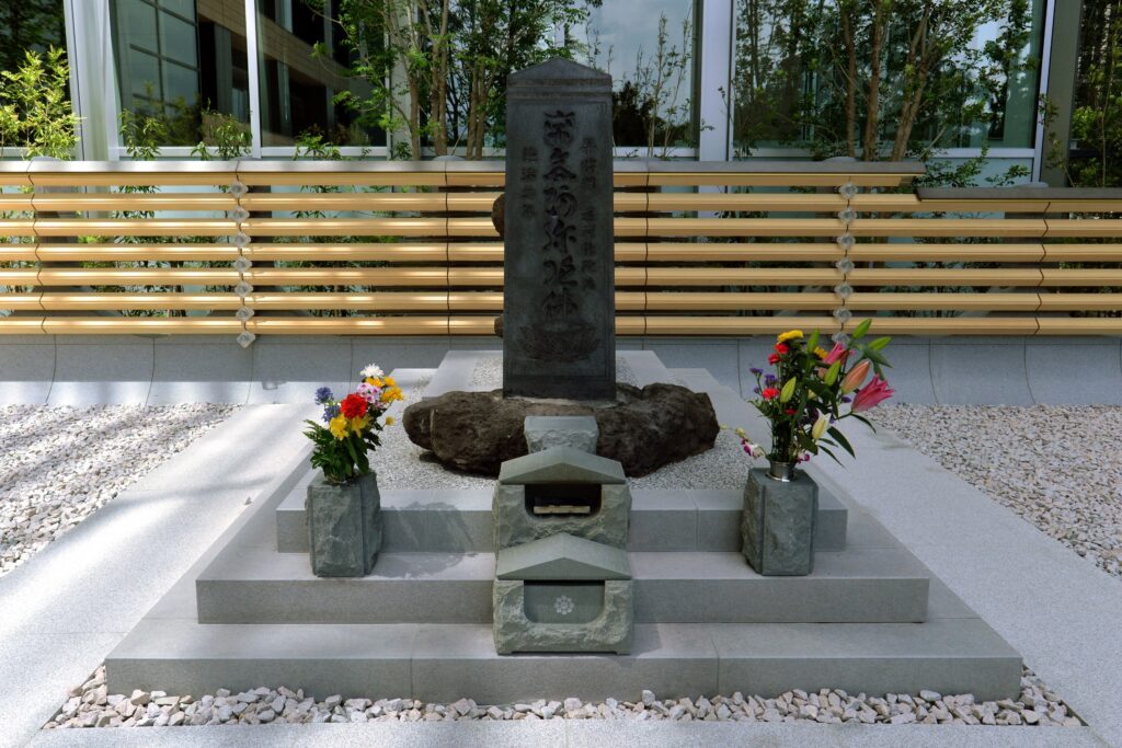 Masakado no kubizuka: stelă de piatră gravată cu caractere japoneze, cu flori proaspete, între clădiri moderne de birouri din Tōkyō